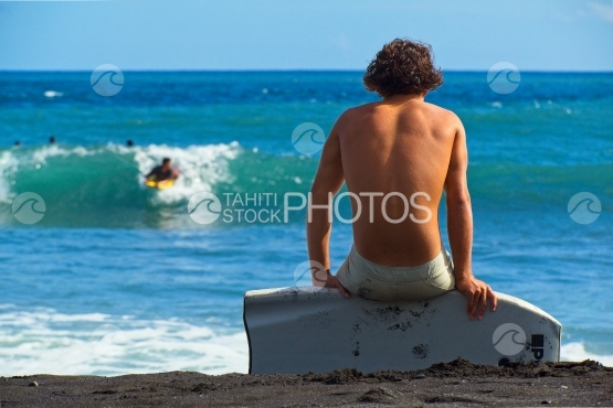 Tahiti, jeune hoomme assis sur son surf sur la plage, surfer dans la vague