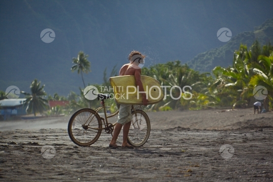 Tahiti, jeune surfer marchant sur la plage de sable noir, avec sa bicyclette